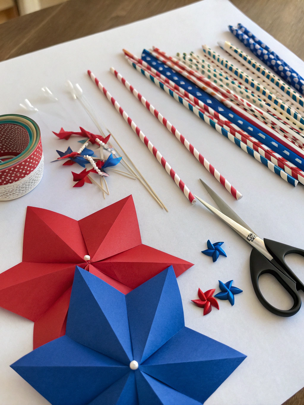 Assortment of craft supplies including red, white, and blue paper, scissors, glue, brads, and wooden dowels laid out on a table.